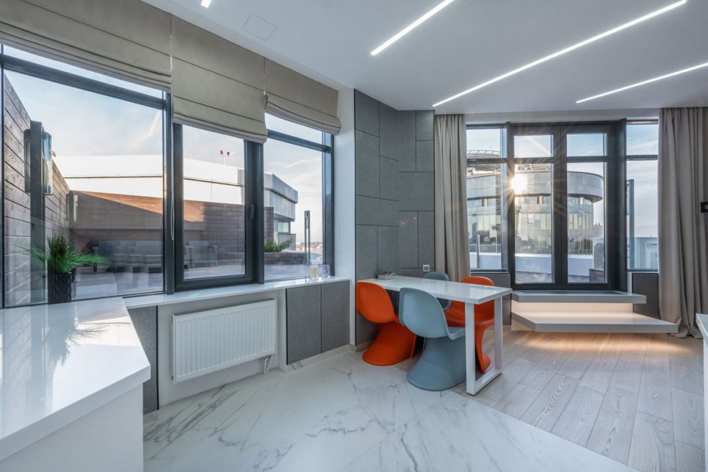 Bright empty spacious studio with windows near jalousie and curtains near table with chairs and counter in sunny day near terrace and buildings