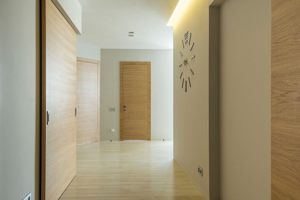 A clean, minimalist corridor featuring wooden doors and floors, with a stylish wall clock.