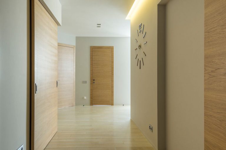 A clean, minimalist corridor featuring wooden doors and floors, with a stylish wall clock.